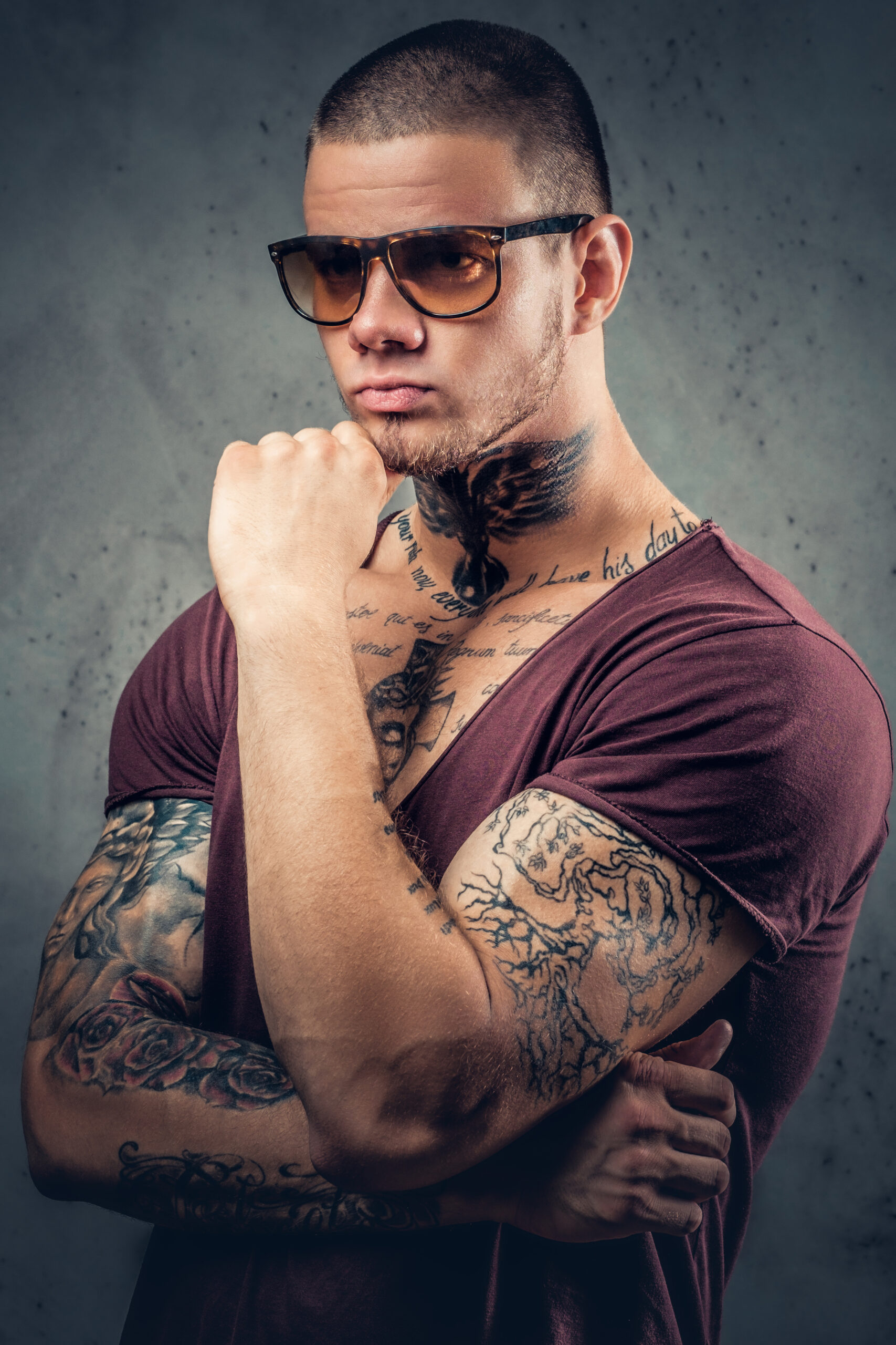 Handsome, athletic male in sunglasses with tattoos on his arms and neck posing over grey artistic background in a studio.